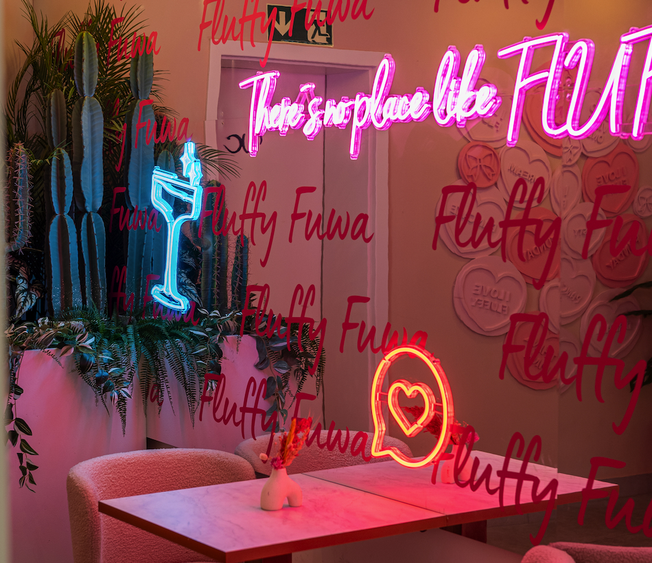 Fluffy Fuwa - IN München - Das Stadtmagazin Cafe interior with pink neon sign and blue cactus-themed neon, glass wall covered in red 'Fluffy Fuwa' text and greenery on a marble table surface