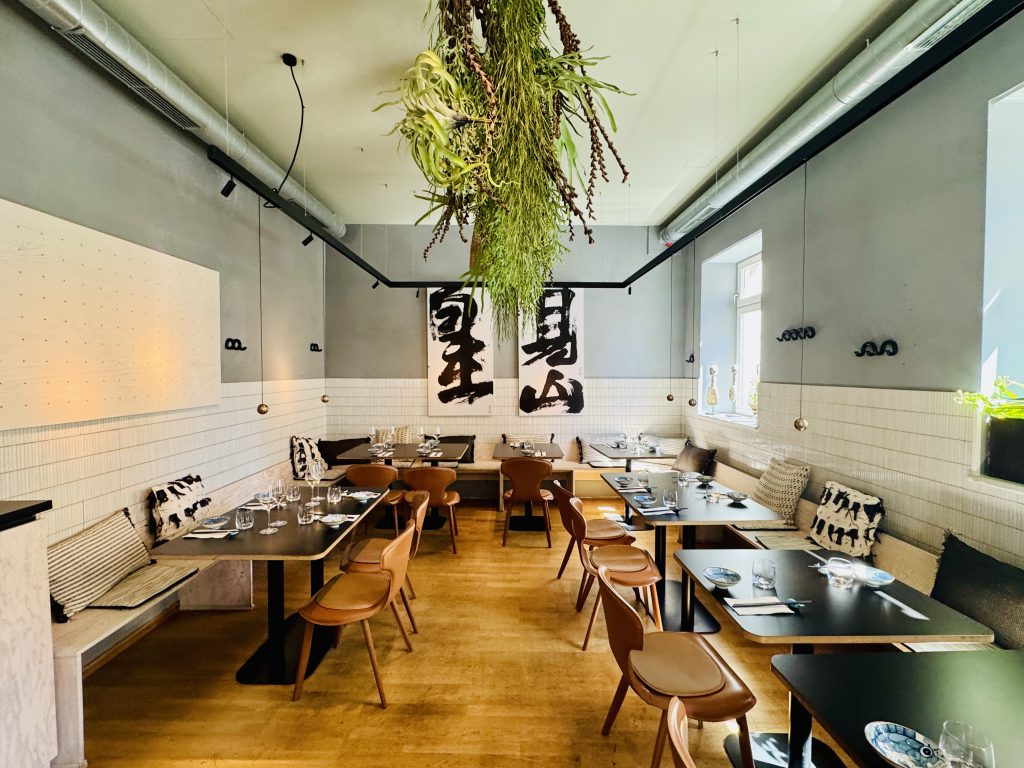 Restaurant dining room with hanging plants from the ceiling, wooden chairs, black tables, and large calligraphy panels on the wall near a tiled backsplash, bathed in natural light from windows.