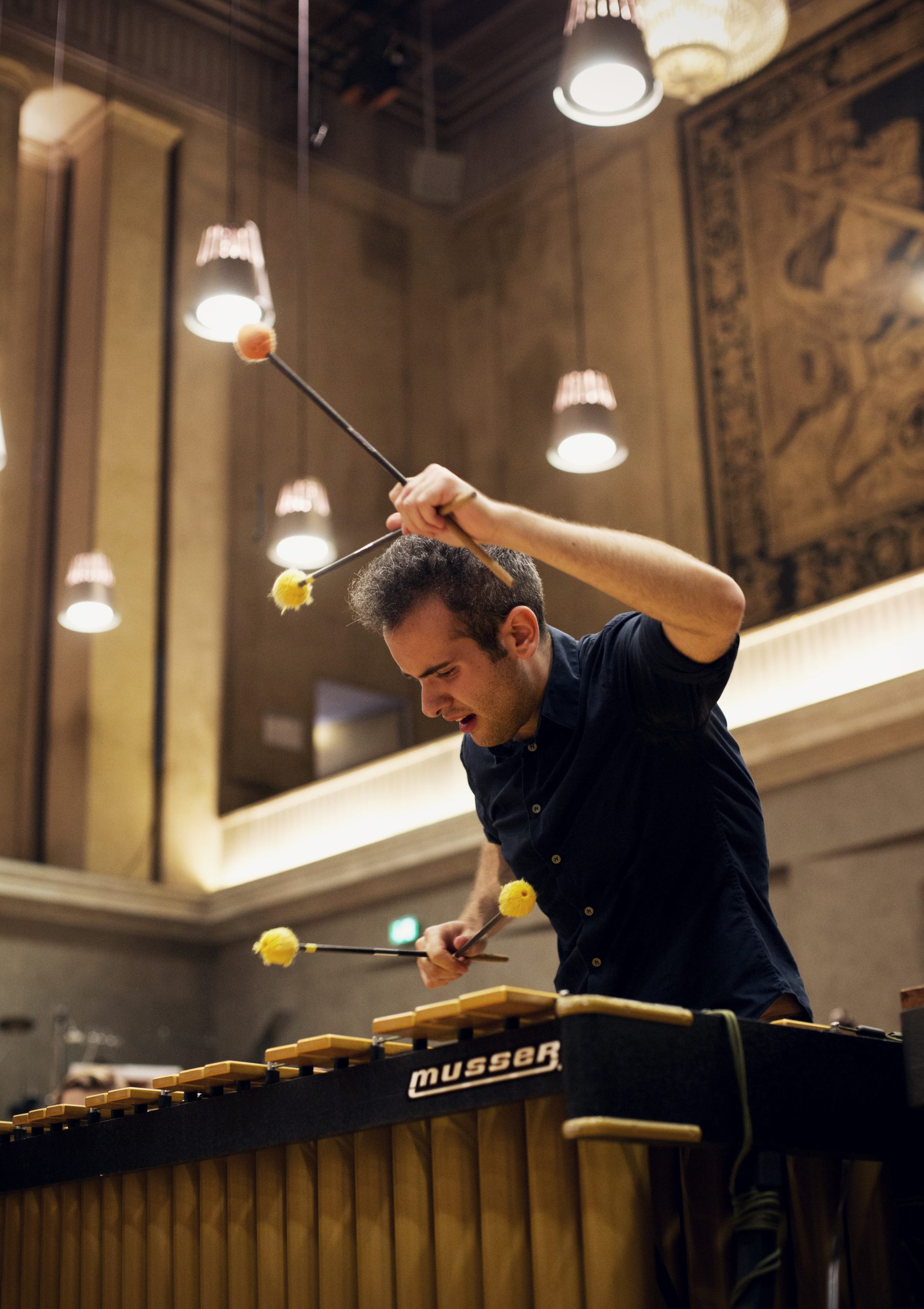 Male musician playing a marimba with yellow mallets in a warmly lit concert hall.