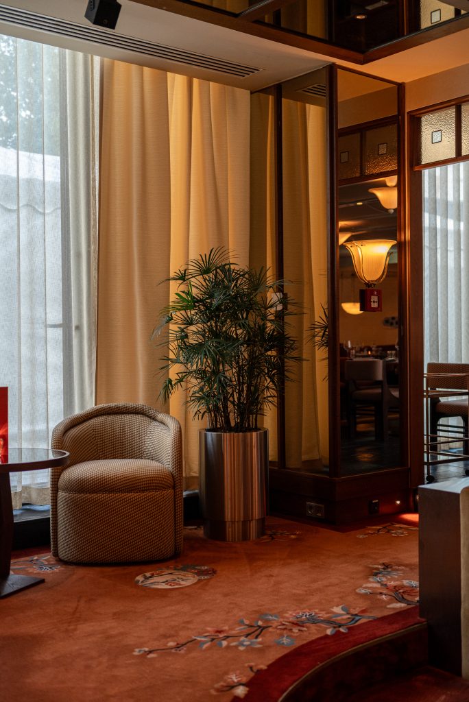 Ibasho-11 - IN München - Das Stadtmagazin Interior hotel lounge with beige curtains, a tall potted plant, and a glass-door reflection showing warm lamps and seating area.