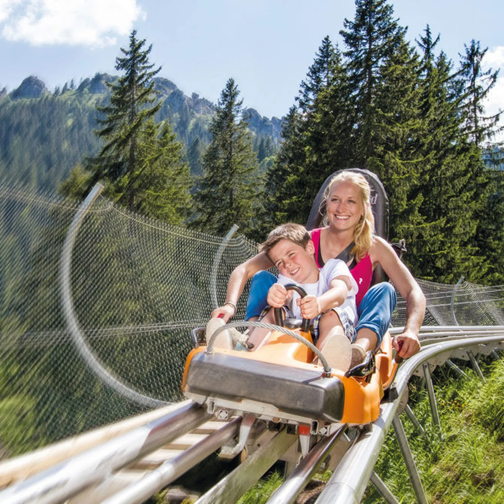 Kolbensattel-Alpine-Coaster-1 - IN München - Das Stadtmagazin Woman and boy smiling in an orange roller coaster car speeding along a track through a pine foresty mountainside.