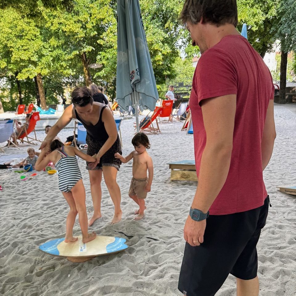 Kulturstrand Balance Boards Workshop - IN München - Das Stadtmagazin Adult guides two young children balancing on a small circular paddleboard on a sandy beach while a man in a red shirt watches nearby.