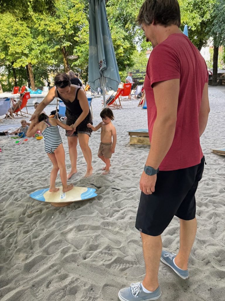 Kulturstrand Balance Boards Workshop - IN München - Das Stadtmagazin Adult guides two young children balancing on a small circular paddleboard on a sandy beach while a man in a red shirt watches nearby.