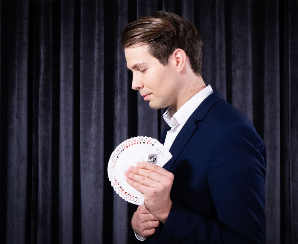 Magic_Close_Up_Thomas_Braun_Zauberschule_Zauberei_Zaubern_Lernen_Bicycle_Karten_Seite - IN München - Das Stadtmagazin Man in a navy suit performing a card trick, holding a fan of playing cards against a dark curtain backdrop.