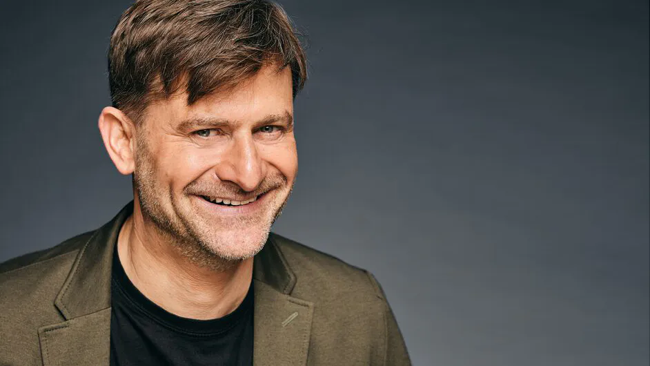 Michael_Altinger_Interview_InMuenchen - IN München - Das Stadtmagazin Smiling man in an olive blazer and black shirt against a dark gray backdrop.