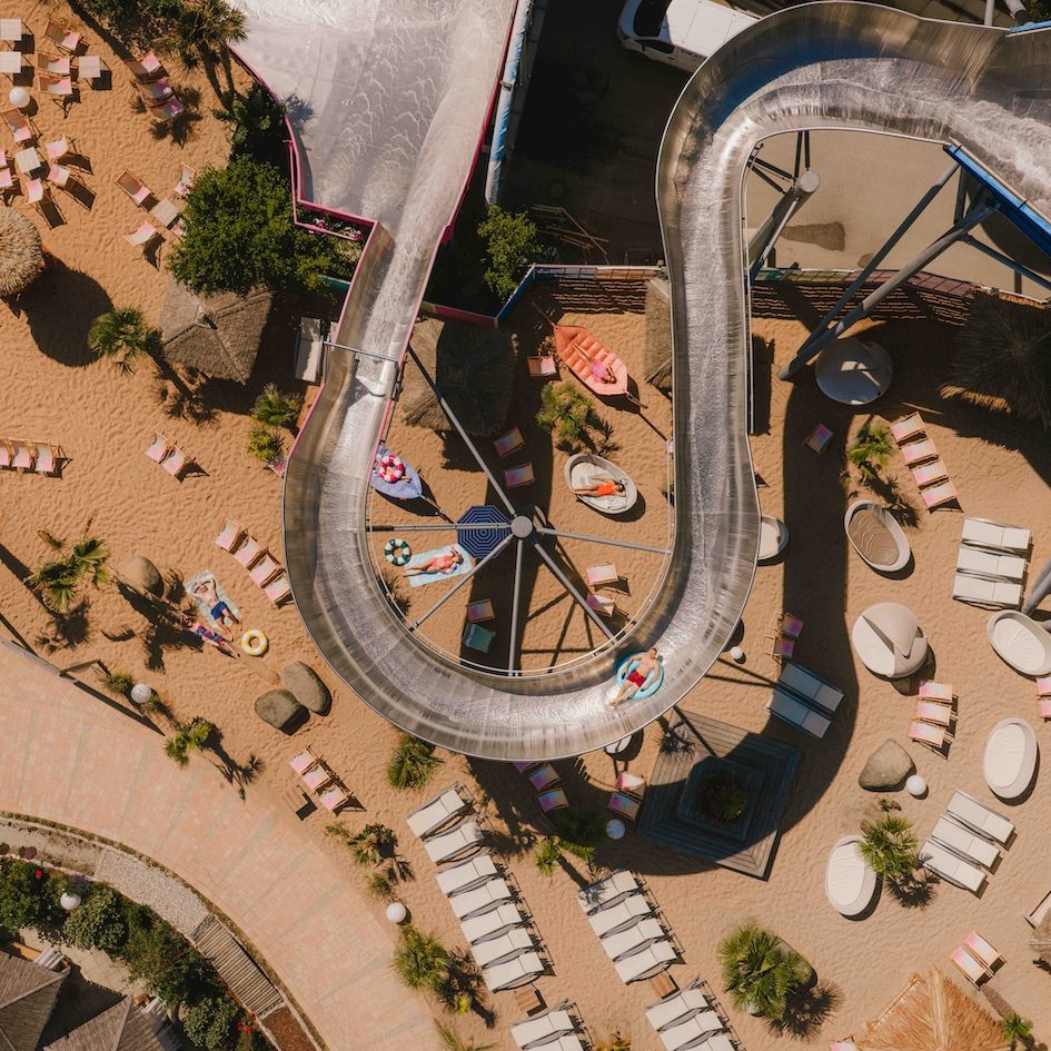 default - IN München - Das Stadtmagazin Aerial view of a winding water slide at a sunny resort, with lounge chairs, palm trees, and a sandy pool area below.