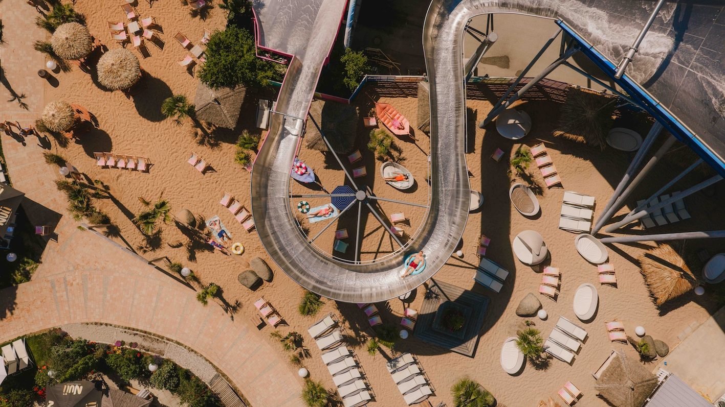 default - IN München - Das Stadtmagazin Aerial view of a twisting water slide spiraling around a central platform, with sun loungers and palm trees on a sandy pool deck.