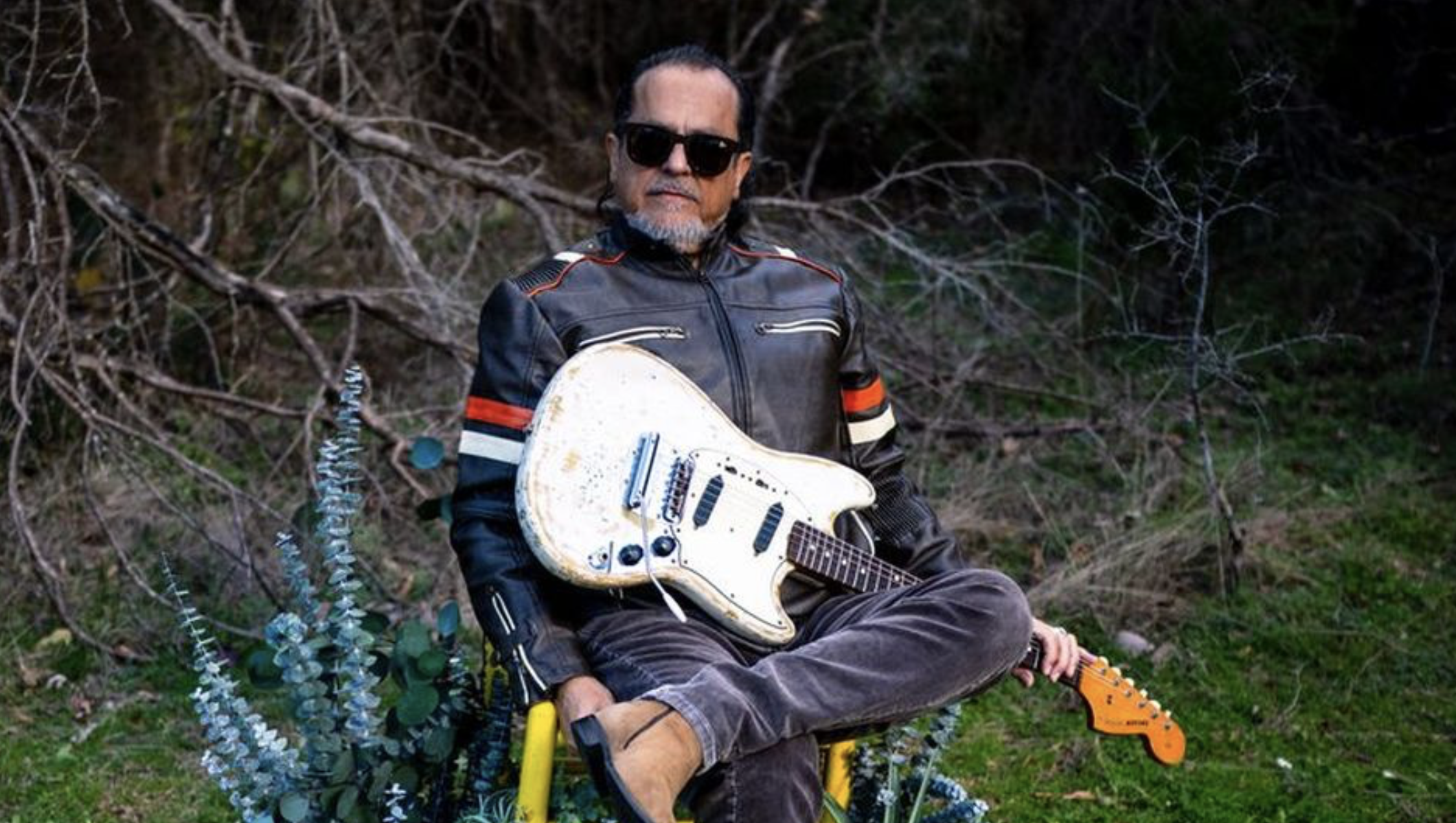 Man wearing a black leather jacket and sunglasses sits outdoors, holding a white electric guitar in a forested area.