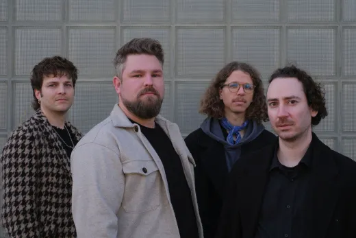 Four men standing side by side in coats in front of a gray glass block wall, looking at the camera.