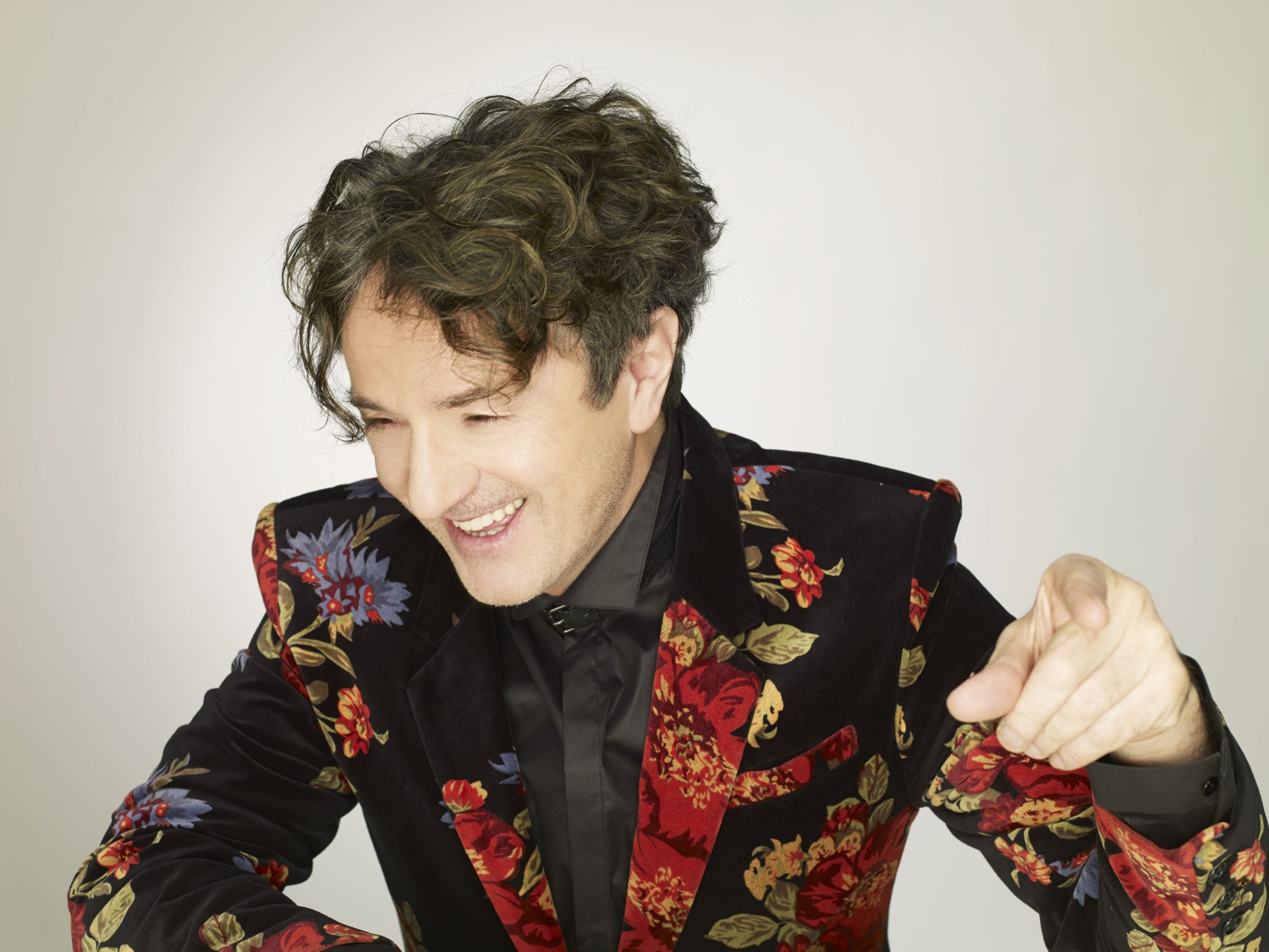 Man in a dark velvet blazer with red and gold floral pattern smiles while leaning forward and extending his hand toward the camera.