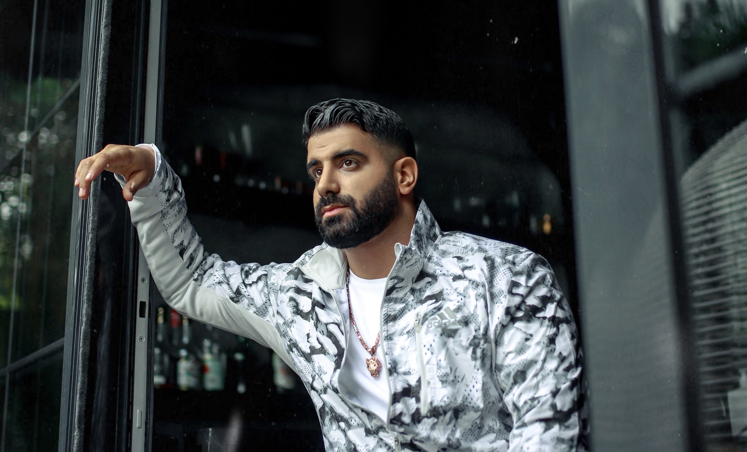 Man with a beard in a white and gray patterned jacket stands in an open doorway, looking to the side.