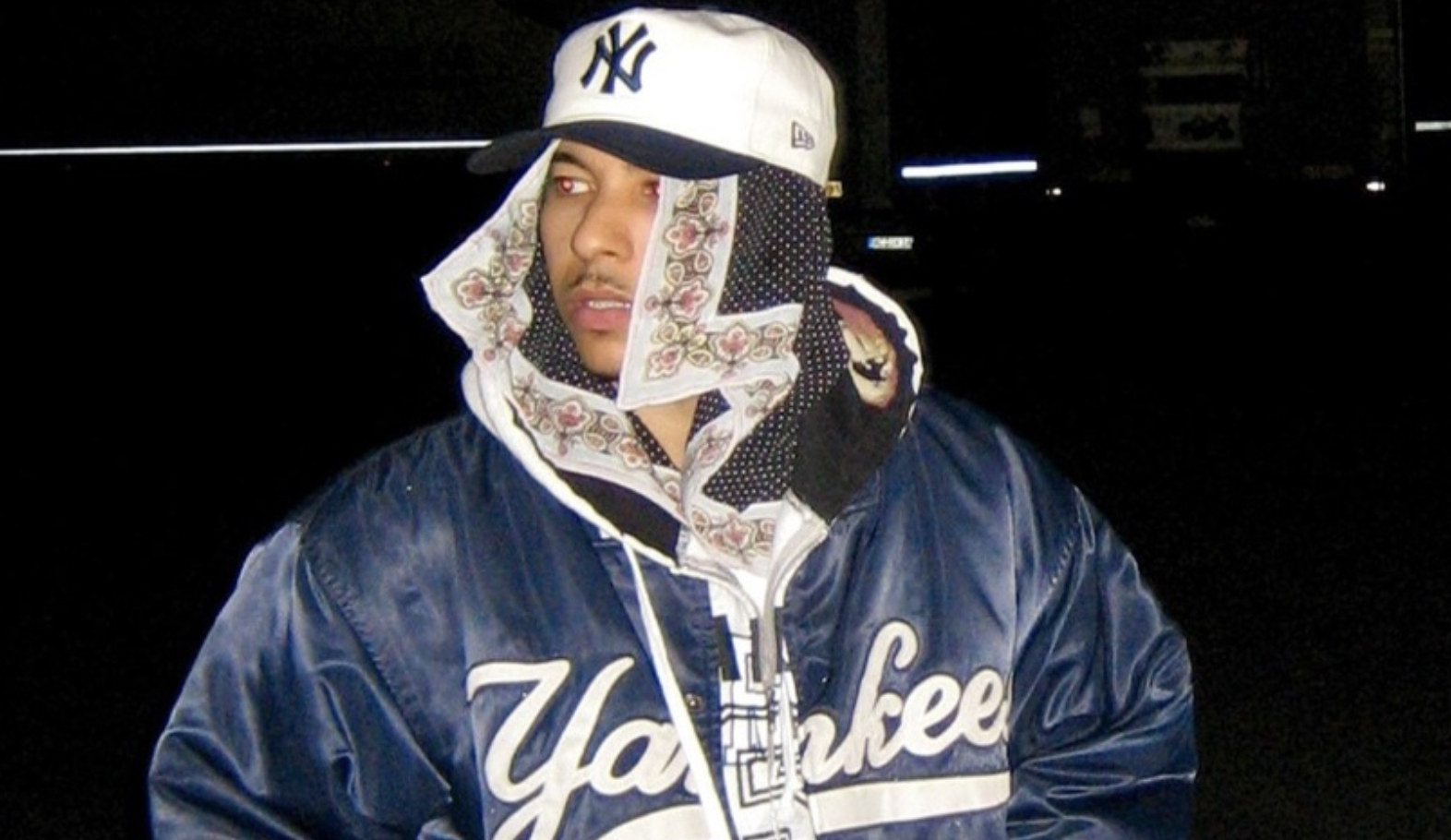 Man wearing a white New York Yankees cap and a patterned scarf wrapped around his face, in a dark nighttime setting with a blue satin Yankees jacket visible.