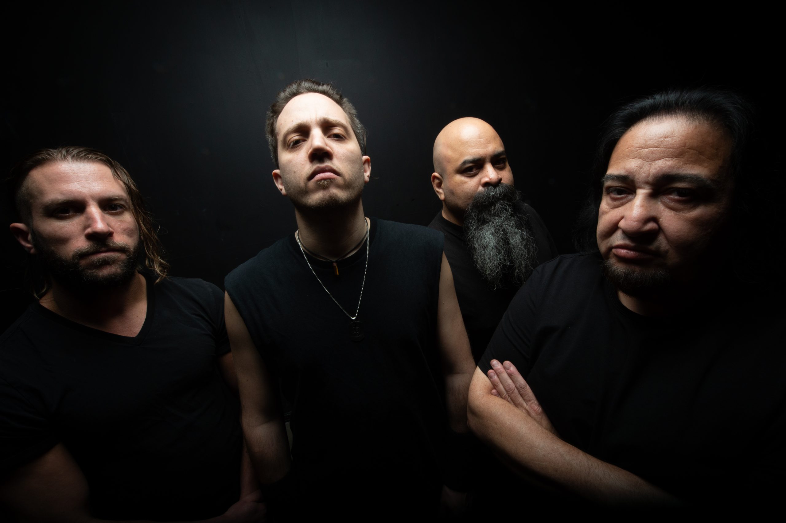 Four men wearing black shirts pose against a dark backdrop, facing the camera with serious expressions.