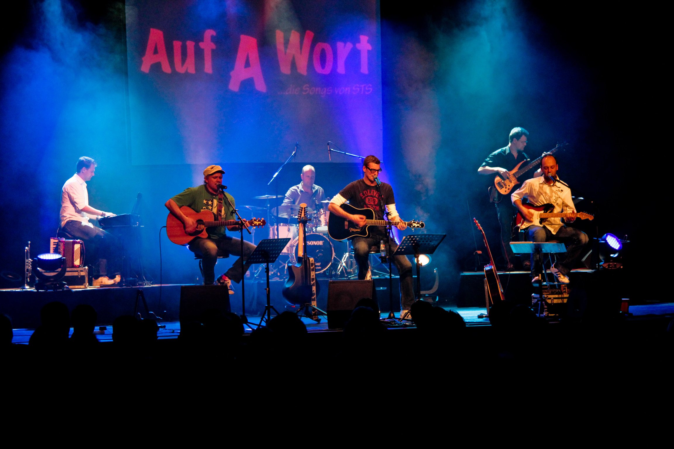 Band on stage performing live with multiple guitarists and a keyboardist, blue and purple stage lights, audience silhouettes in front, banner with bold red letters in the background.