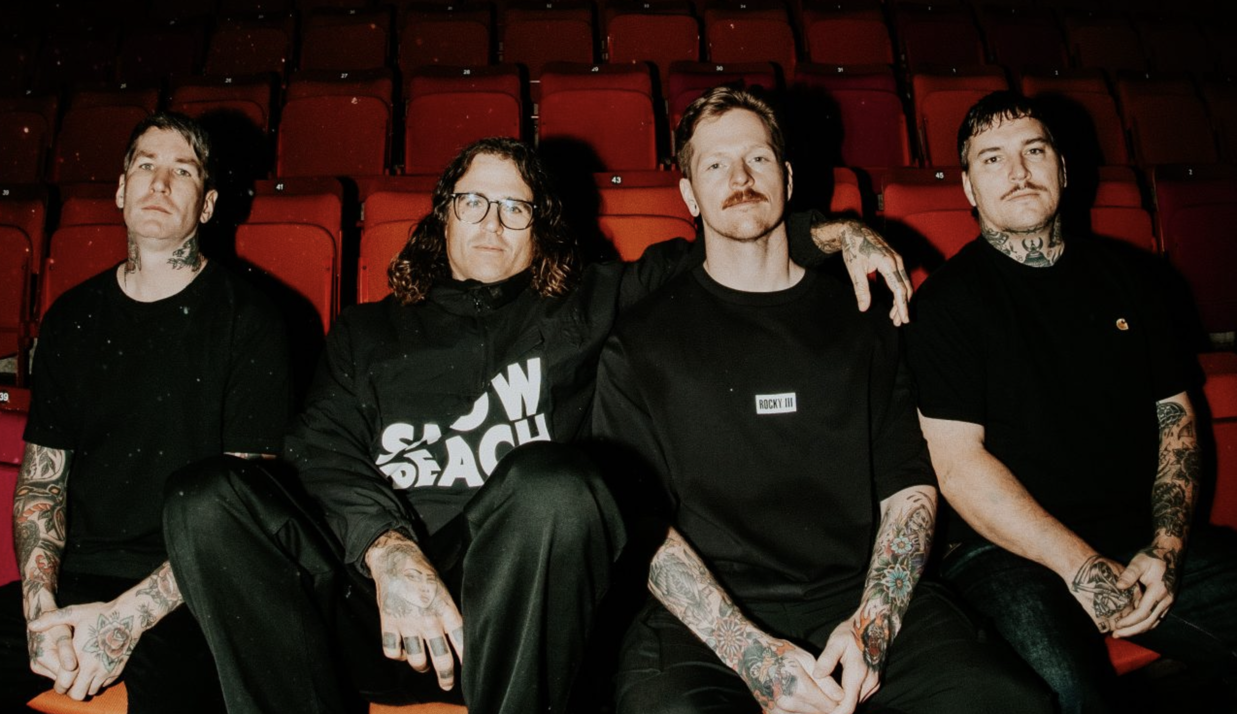 Four tattooed men in black sit together in red theater seats, looking at the camera.