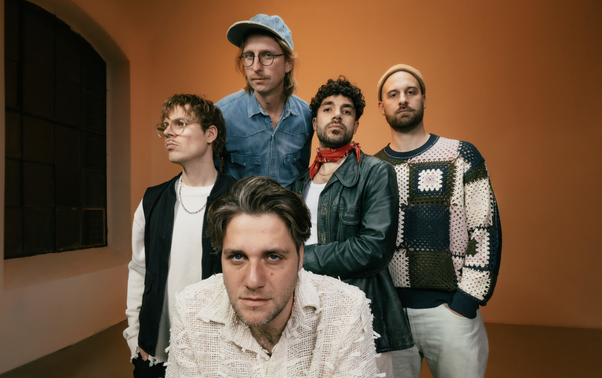 Group portrait of four men posing against an orange studio wall; front man wears a cream crocheted sweater, others in denim, leather, and a patterned knit.