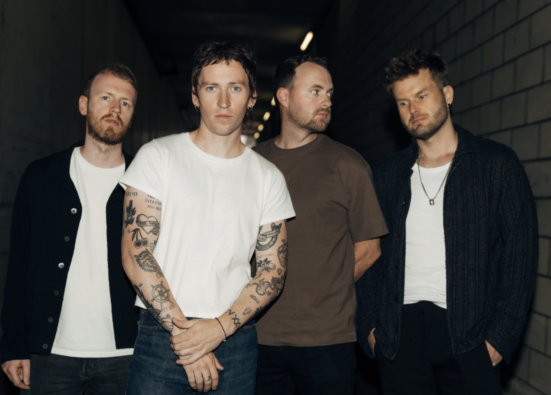 Four men standing together in a dim hallway, casual outfits; the man in a white T-shirt shows tattoos on his arms in the foreground.