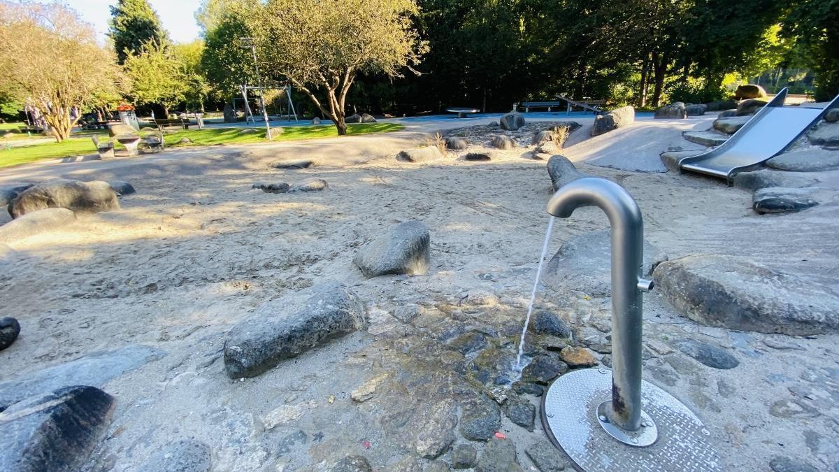 Public park playground with a metal water fountain in the foreground, a sandy play area, rocks, and a slide in the background.