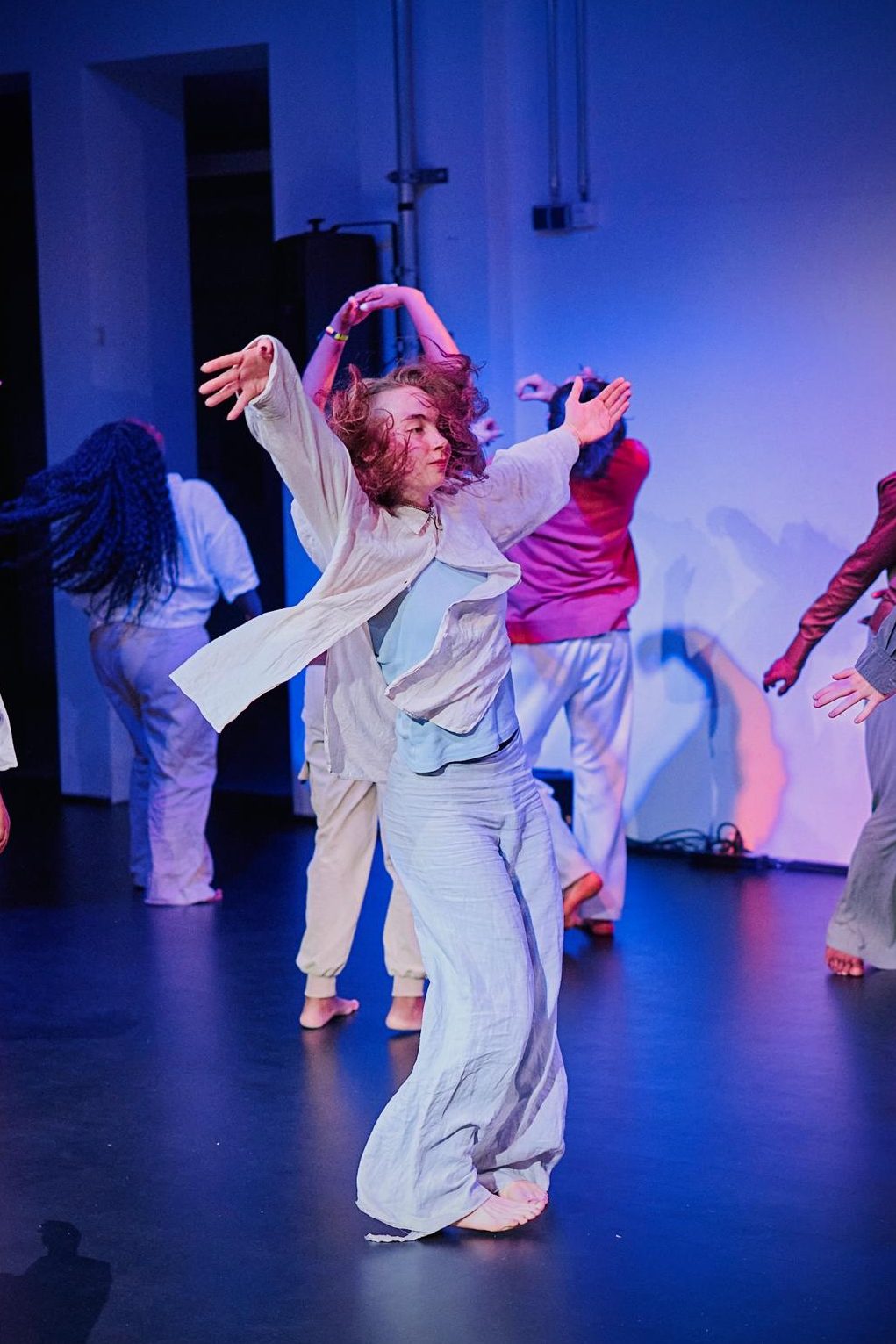 ZiK_c_Philip Nemenz - IN München - Das Stadtmagazin Dancer with arms raised in a light blue outfit, barefoot on a dark stage under blue lighting; other dancers in the background.