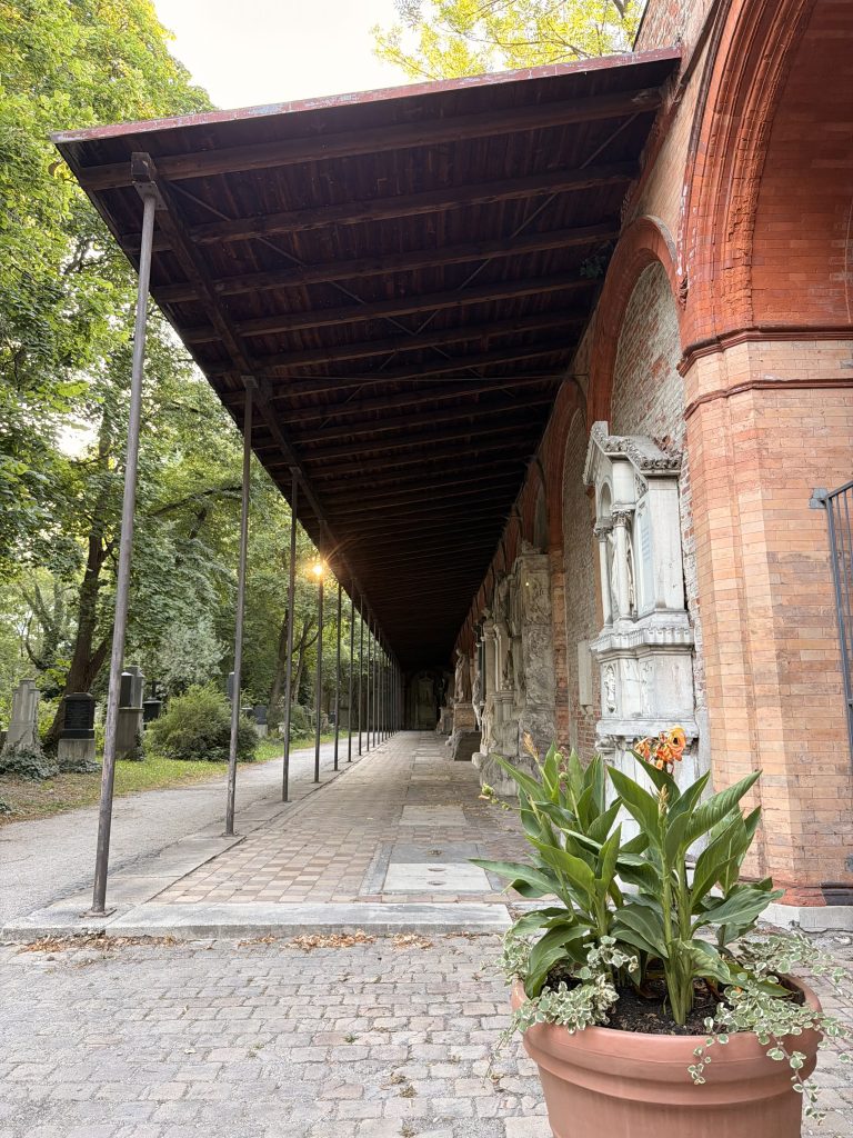 Brick arches and an overhanging wooden roof line a long walkway beside a garden path; a large potted plant sits in the foreground.