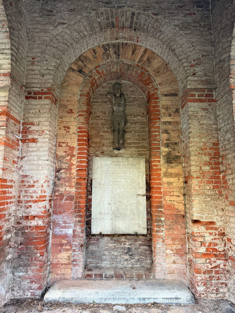 Weathered brick memorial alcove with a stone armored statue centered under arched brickwork, and a plaque below.