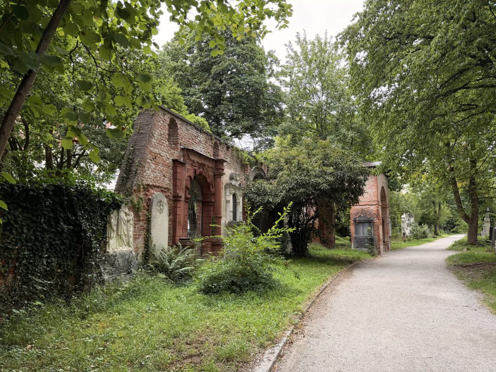 Brick-red brick wall with arched openings and carved statue niche in a leafy cemetery, a gravel path curving beside it.