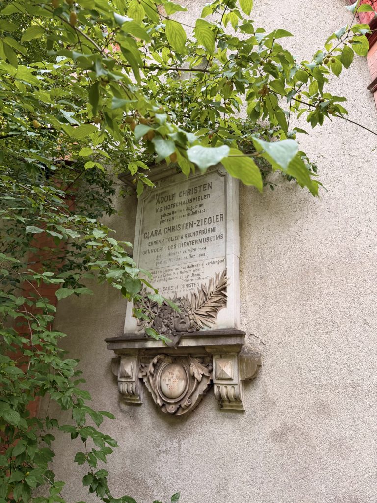 Stone memorial plaque mounted on a weathered wall, largely obscured by green vines and leaves.