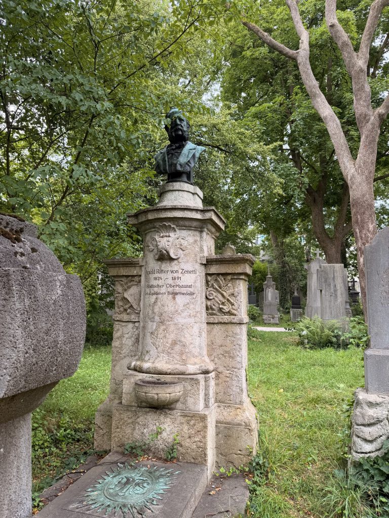 Bronze bust of Arnold Ritter von Zenetti on a carved stone pedestal in a leafy cemetery with other gravestones nearby.