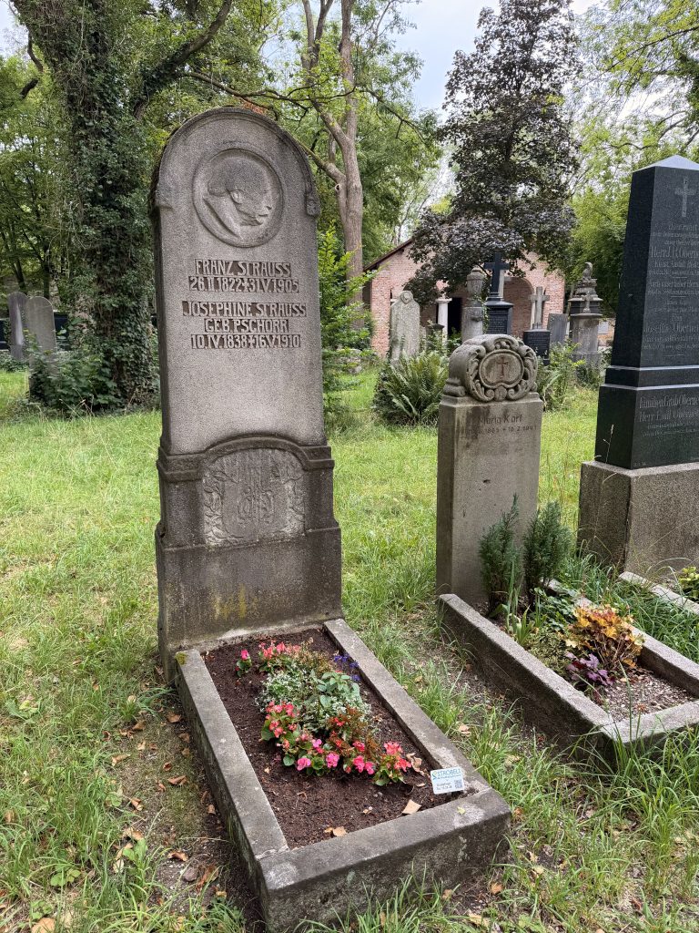 Weathered cemetery headstone surrounded by trees, with the name Franz Strauss carved on it and a small flower bed nearby.