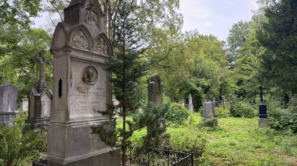 Large stone tomb with carved reliefs and a cross, surrounded by an iron railing in a leafy cemetery.