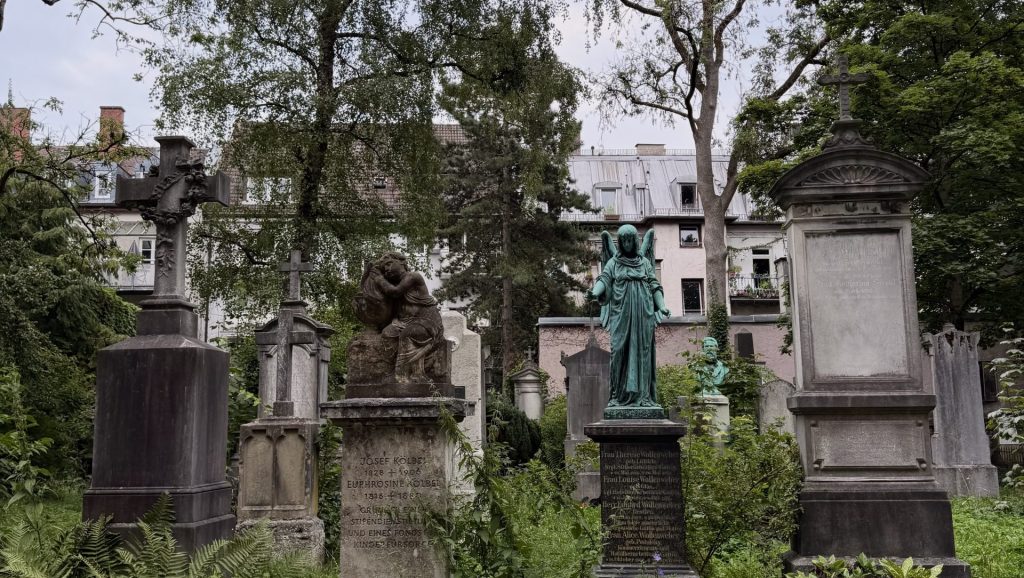 Overgrown cemetery with weathered headstones, a green statue of an angel, and dense ferns in the foreground. Beneath tall trees and a muted city backdrop.