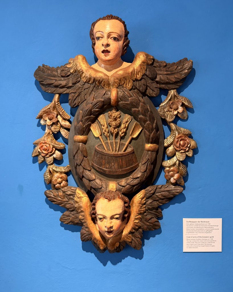 biermuseum-Muenchen-zunftwappen-bierbrauer - IN München - Das Stadtmagazin Aufwendiges Zunftwappen aus Holz, welches Engel zeigt, die ihre Flügel um die Zutaten des Biers entfalten im Biermuseum München