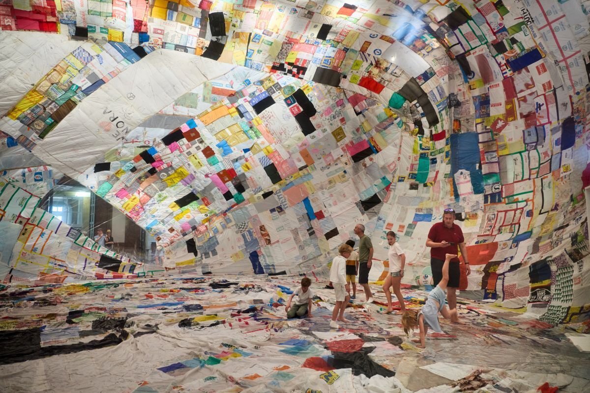 Colorful fabric quilt interior shaped as a tunnel, with people—children and adults—walking and playing on the patchwork floor and walls.