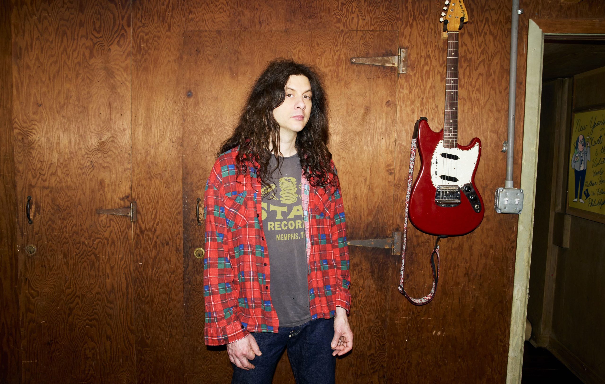 kurt vile - IN München - Das Stadtmagazin Person with long dark hair wearing a red plaid shirt over a gray graphic tee stands against a wooden wall beside a red electric guitar hanging on the wall.