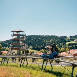 sommerrodelbahn-bayerischer-wald-coaster-2 - IN München - Das Stadtmagazin Person seated on a wooden roller coaster track in a sunny outdoor amusement park, with a tall spiral tower and green hills in the background.