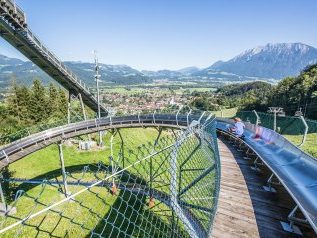 sommerrodelbahn-oberaudorf-Chiemsee-Alpenland-Tourismus-1 - IN München - Das Stadtmagazin Coaster track winds around above green fields, with riders visible on the ride and a valley, hills, and distant mountains under a clear blue sky.
