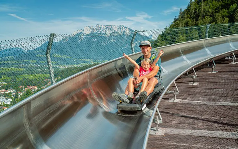 sommerrodelbahn-oberaudorf-Chiemsee-Alpenland-Tourismus-2 - IN München - Das Stadtmagazin A man and a young girl ride a curved metal slide down a hillside, smiling as they descend with a mountain landscape in the background.
