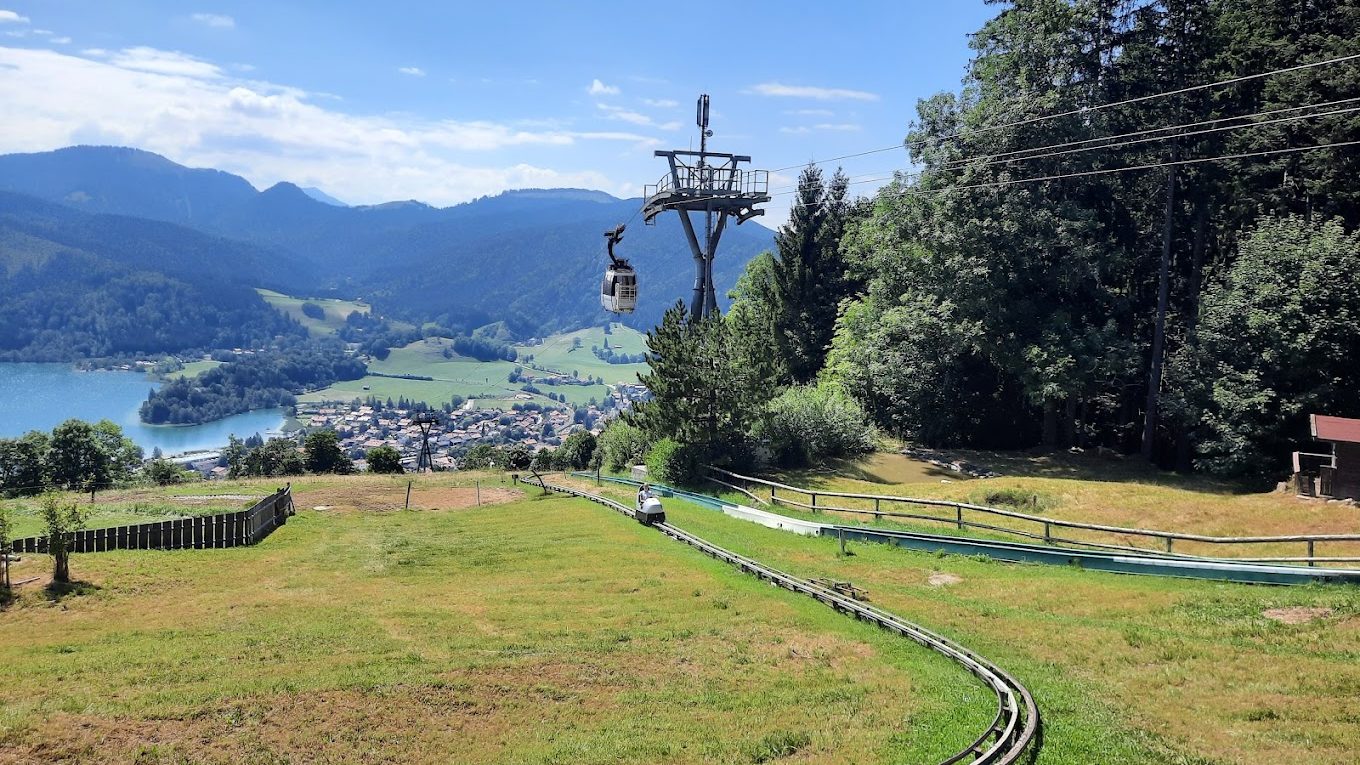 sommerrodelbahn-schliersbergalm - IN München - Das Stadtmagazin Gondola lift car ascending along a hillside with a curved track, overlooking a lake and a village below, surrounded by mountains and trees.