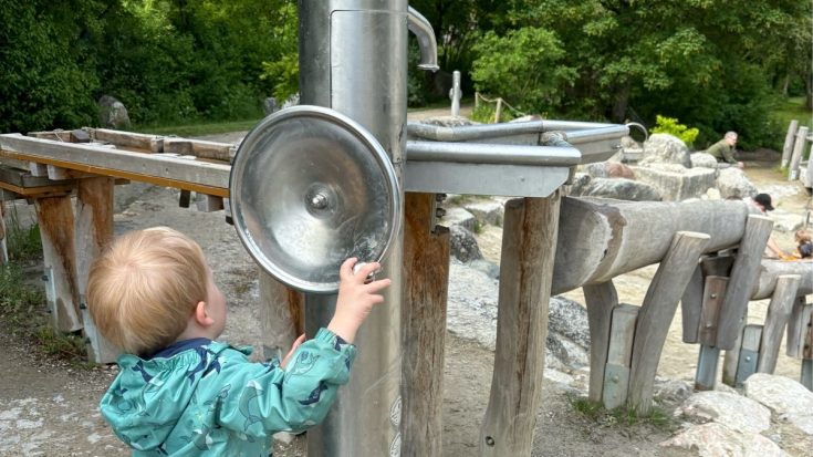 Child in a teal jacket reaches for a large metal hand pump in an outdoor park water feature, with wooden posts and rocks in the background.