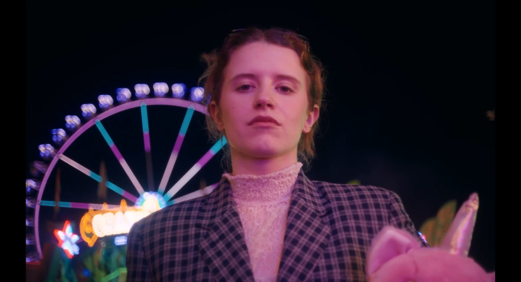 wfilm_werweiss_13 - IN München - Das Stadtmagazin Young person in a plaid blazer poses in front of a colorful, neon-lit Ferris wheel at night, carnival mood.