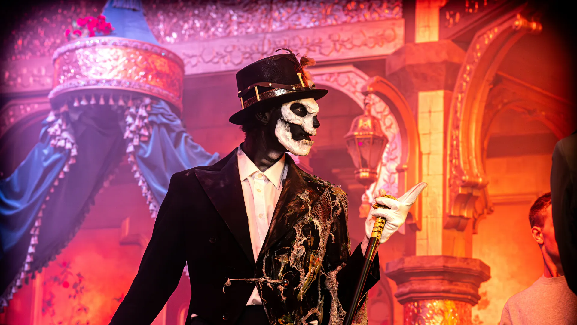 Performer with skull face paint in a black tuxedo and top hat, holding a cane on a vibrant stage with ornate backdrop.