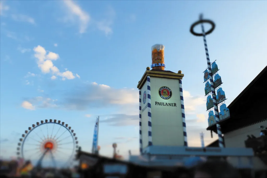 Paulaner - IN München - Das Stadtmagazin Paulaner festival tower with blue-and-white striped pillars and logo, ferris wheel in the background against a blue sky