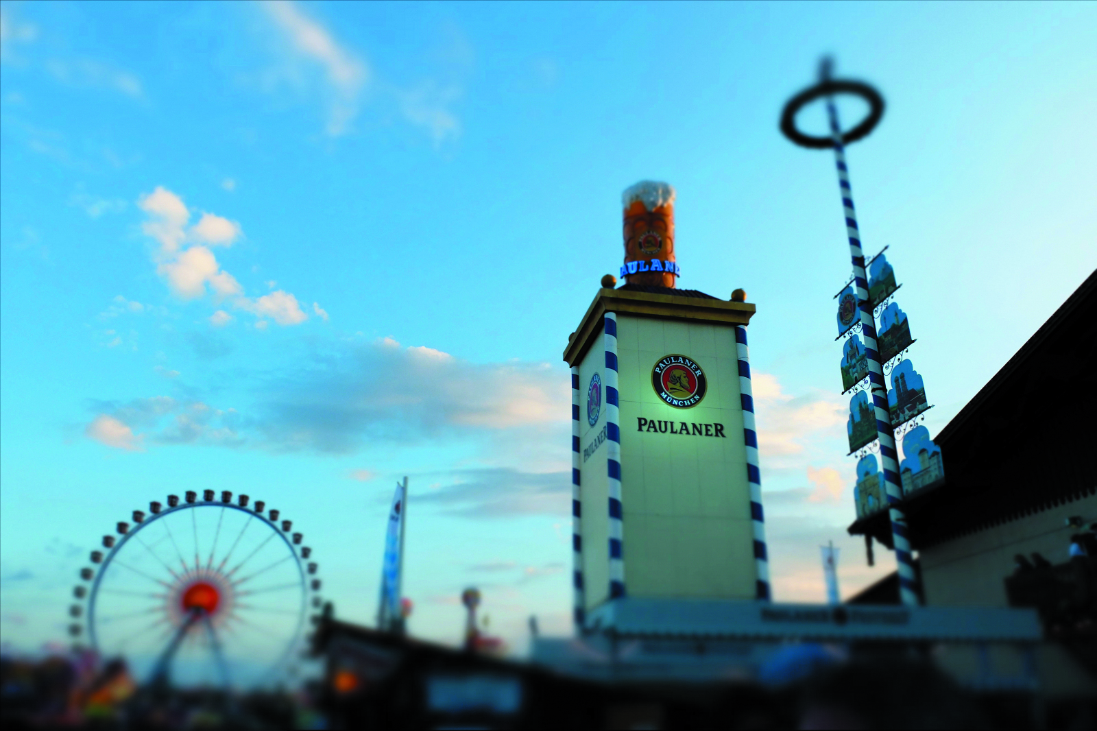 Paulaner festival tower with blue-and-white striped pillars and logo, ferris wheel in the background against a blue sky