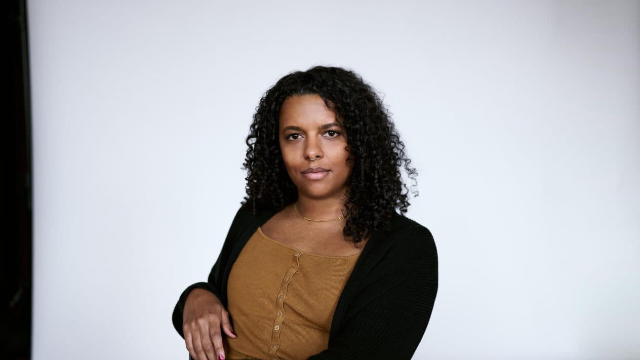 Alice Hasters - IN München - Das Stadtmagazin Portrait of a woman with curly hair, wearing a brown top and black cardigan, standing against a light background.