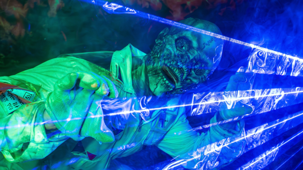 6 - IN München - Das Stadtmagazin Zombie prop in a green shirt presses against a clear plastic barrier bathed in blue light.
