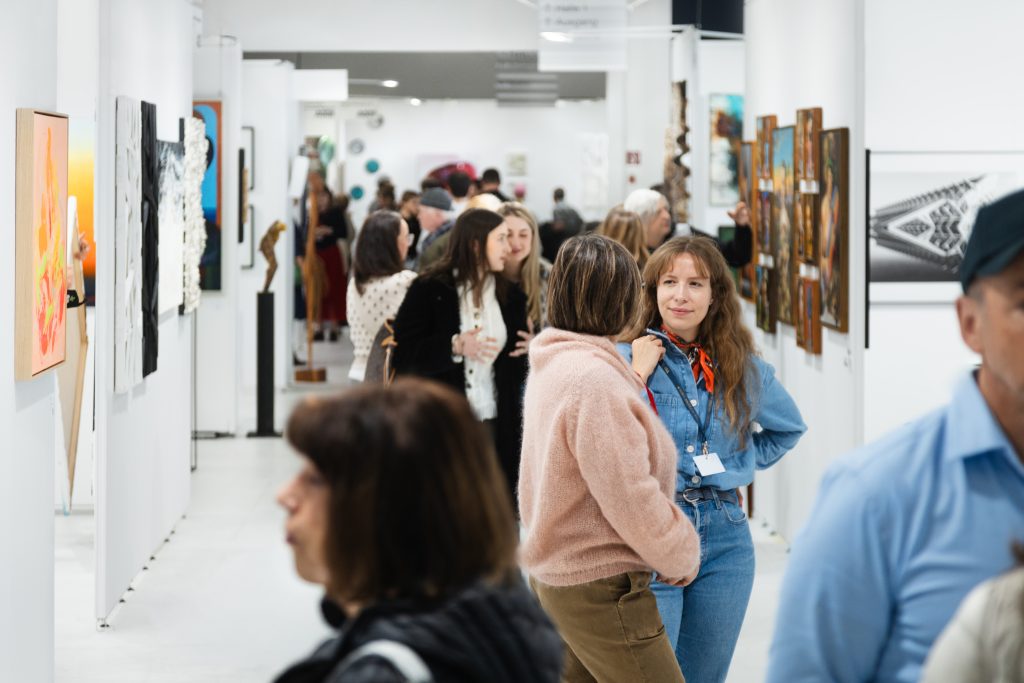 ARTMUC-Kunstmesse_c_by_Isabella_Kilian_Druck_Querformat_No_44 - IN München - Das Stadtmagazin People gather in a bright art gallery viewing framed artworks along white walls.