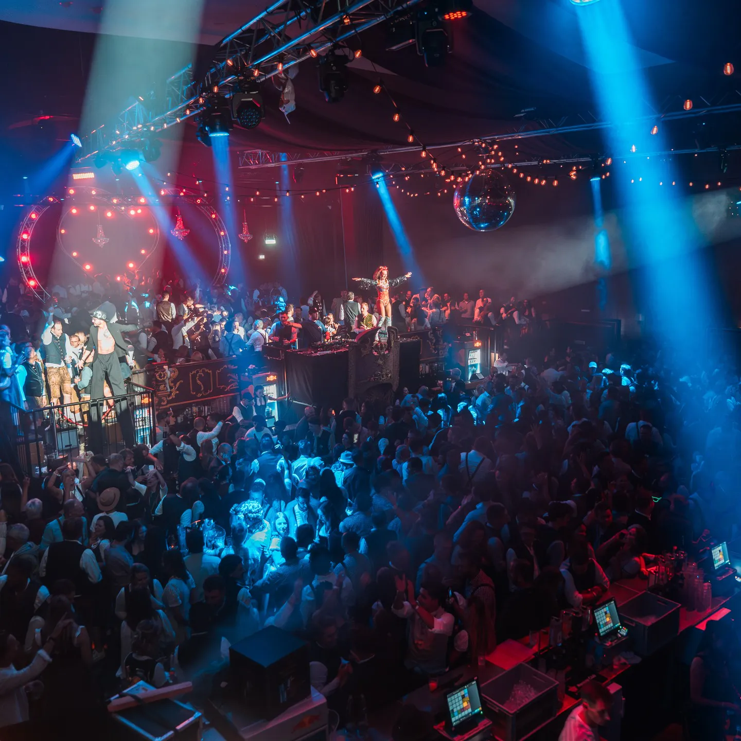 Busy nightclub scene with a dancer on a throne at the DJ booth, crowd dancing under blue and red lights and a hanging disco ball.