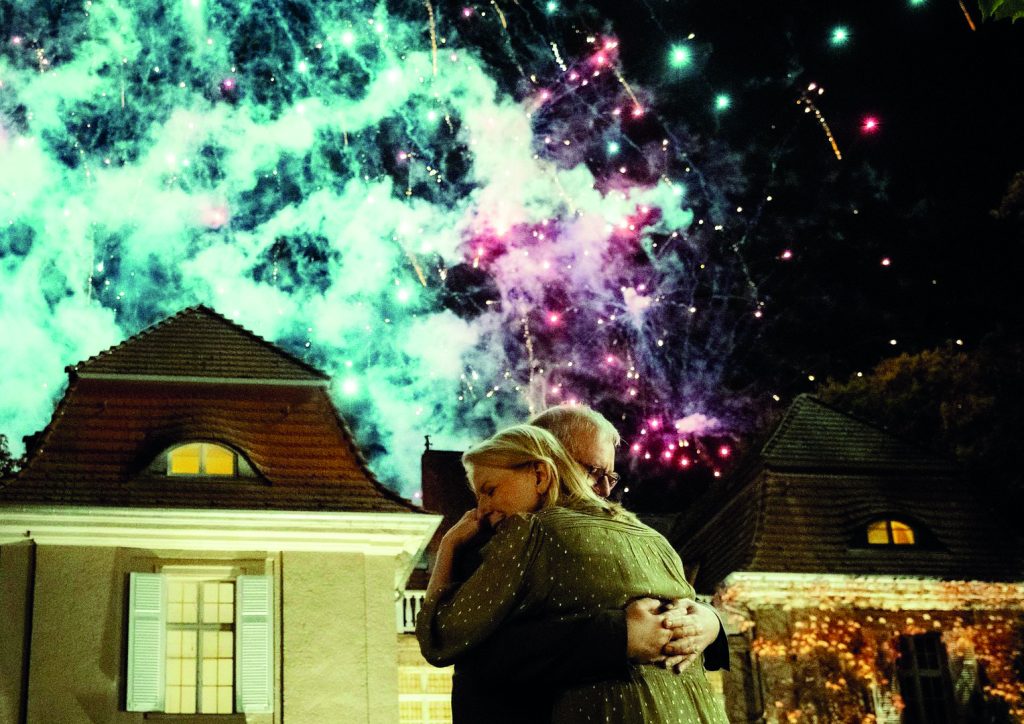 Alte liebe - IN München - Das Stadtmagazin Two people hug in the foreground as green and pink fireworks light the night sky above a rooftop scene.