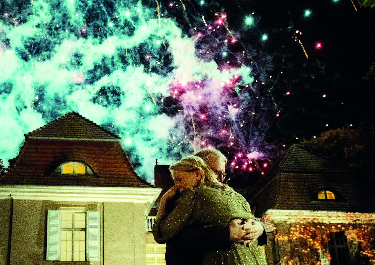 Two people hug in the foreground as green and pink fireworks light the night sky above a rooftop scene.