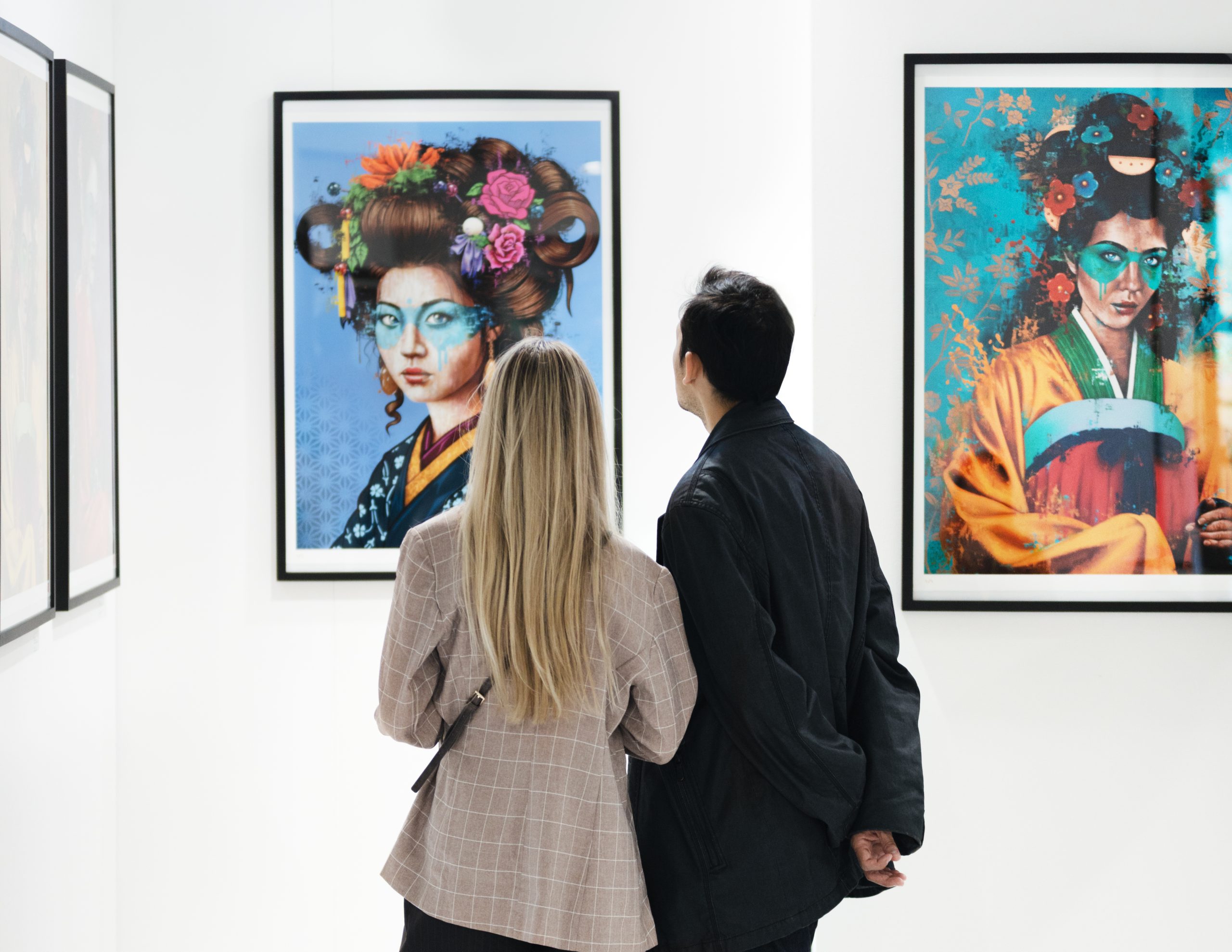 Two visitors in a white-walled gallery gaze at framed, vibrant geisha portraits displayed on the wall from the back.